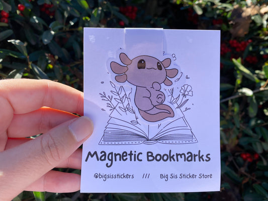 Hand holding a card with an axolotl magnetic bookmark and text 'Magnetic Bookmarks' against a blurred green background.