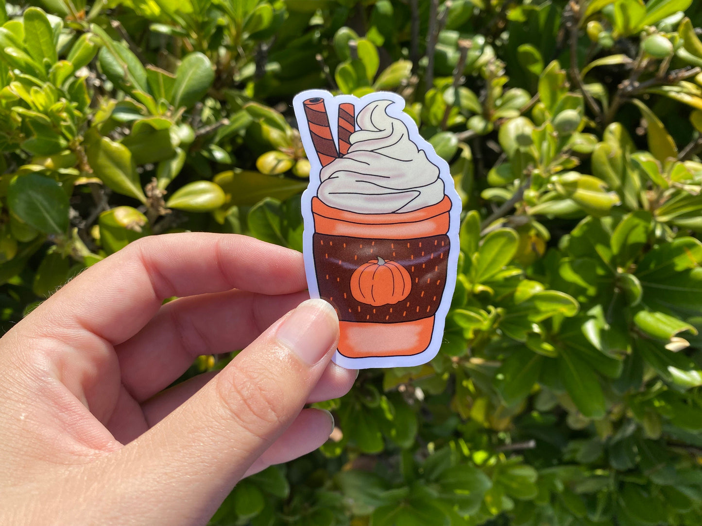 Hand holding a sticker of a cup with whipped cream against a green leafy background
