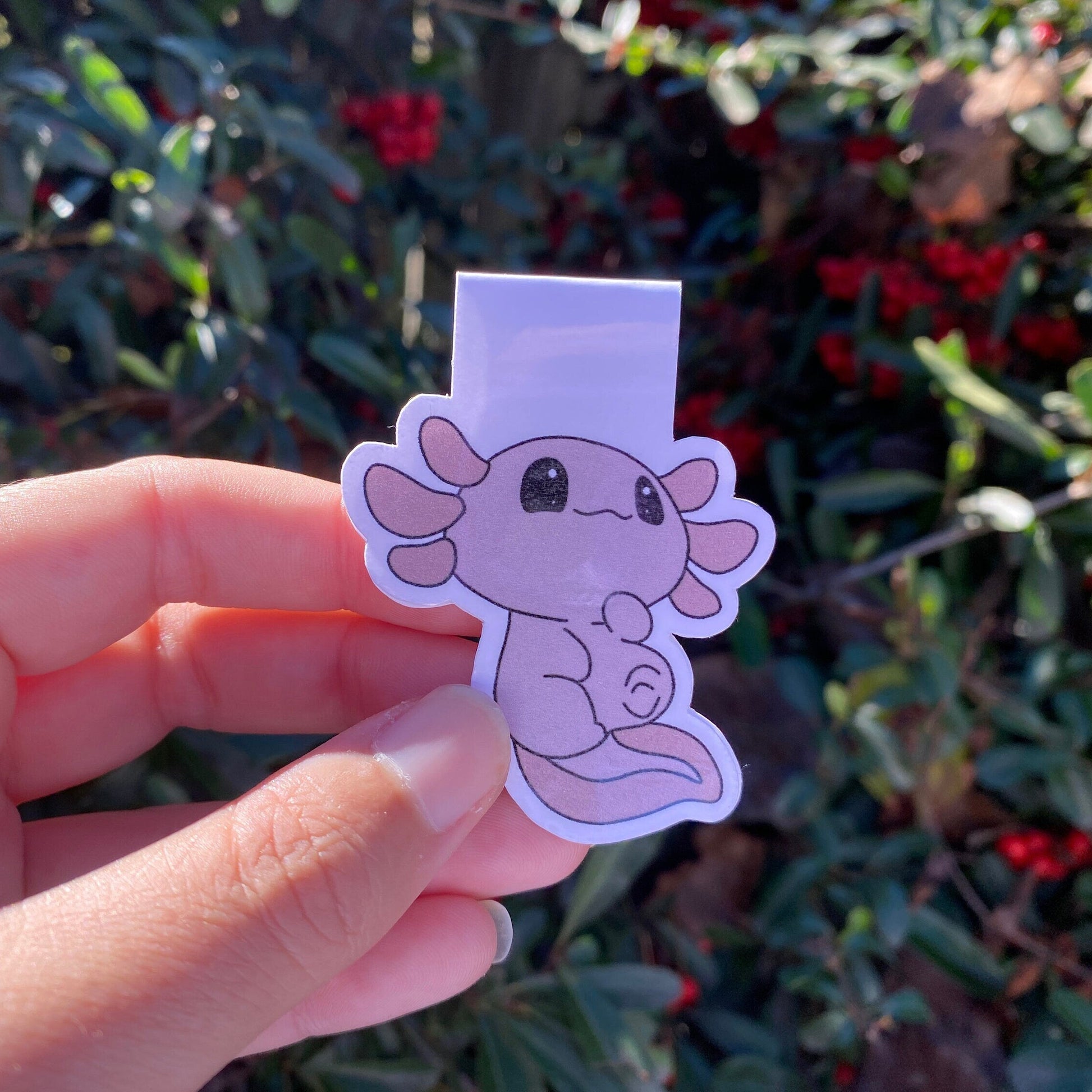 Hand holding a pink axolotl magnetic bookmark in front of green foliage with red berries