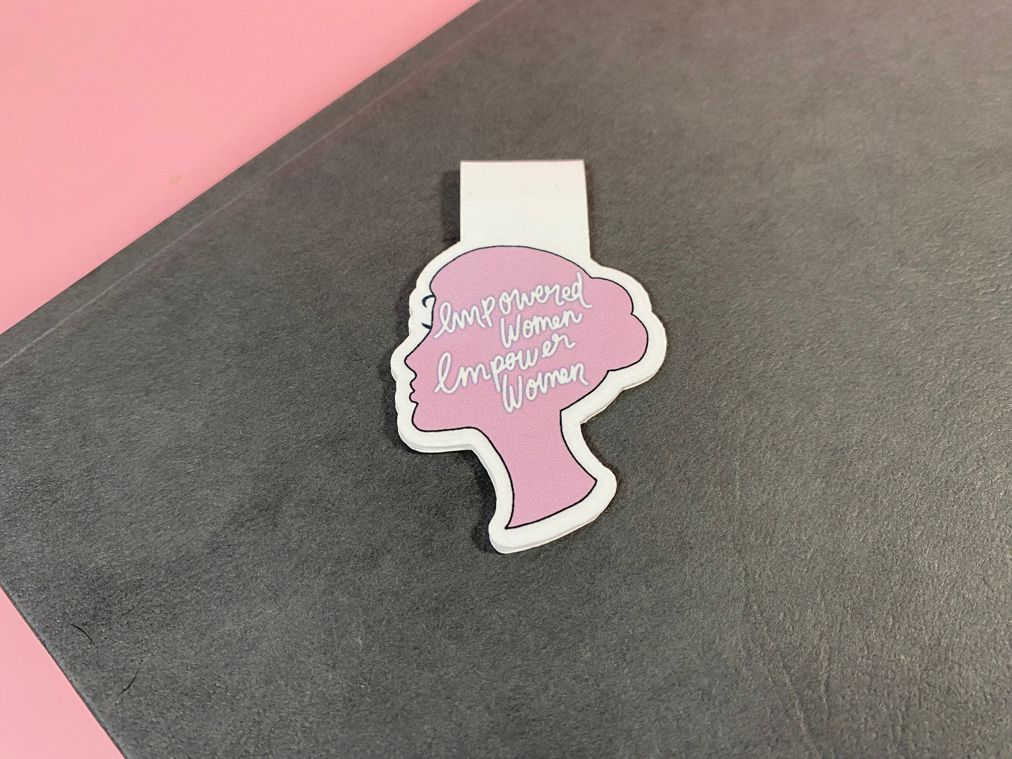 Pink and white magnetic bookmark with a silhouette of a woman's head and 'Empowered Women Empower Women' text on a gray surface.