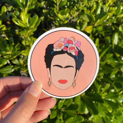 Circular sticker with a stylized illustration of Frida Kahlo held in front of green foliage