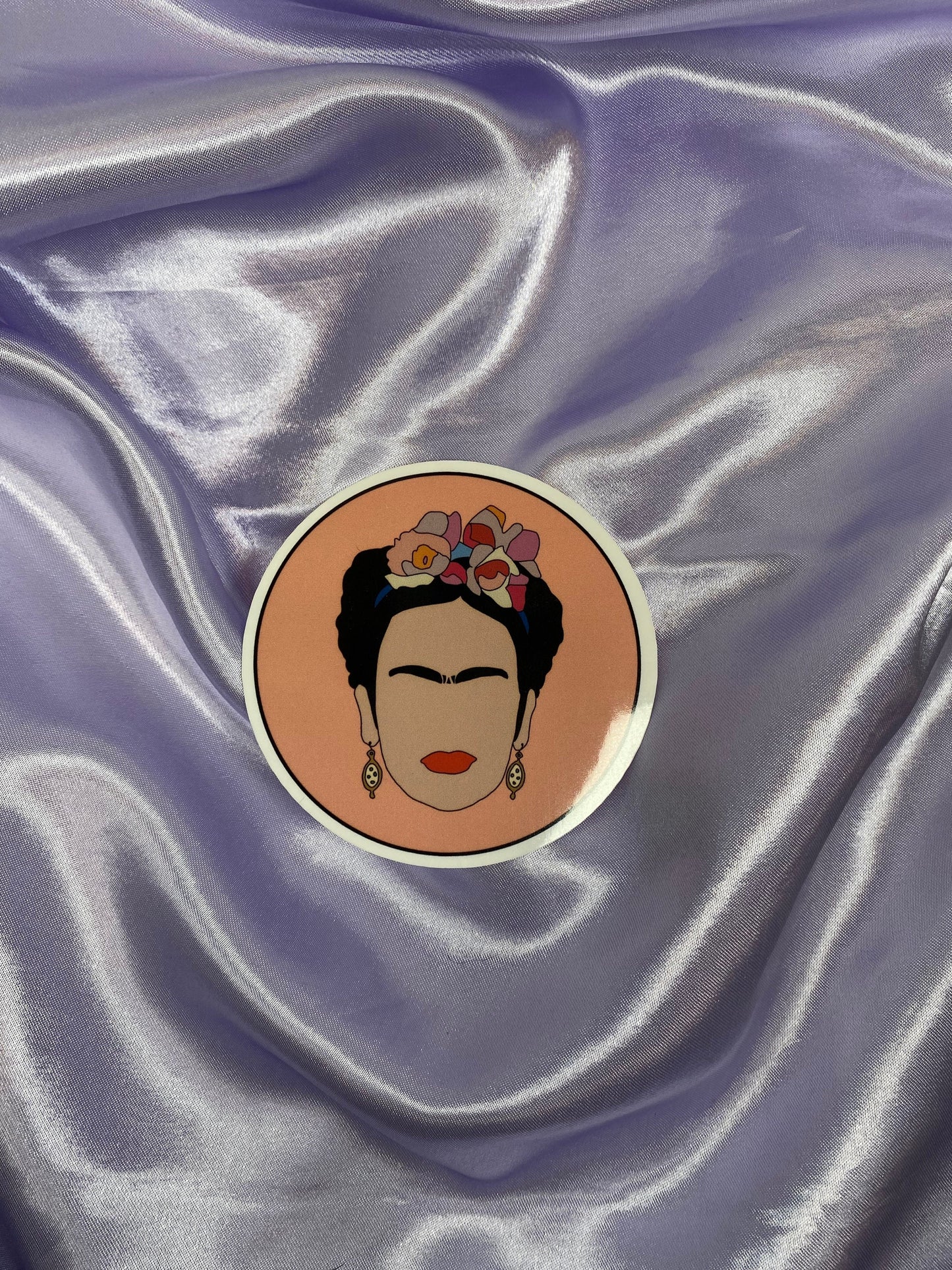 Circular sticker of a stylized portrait of Frida Kahlo on a textured purple fabric background