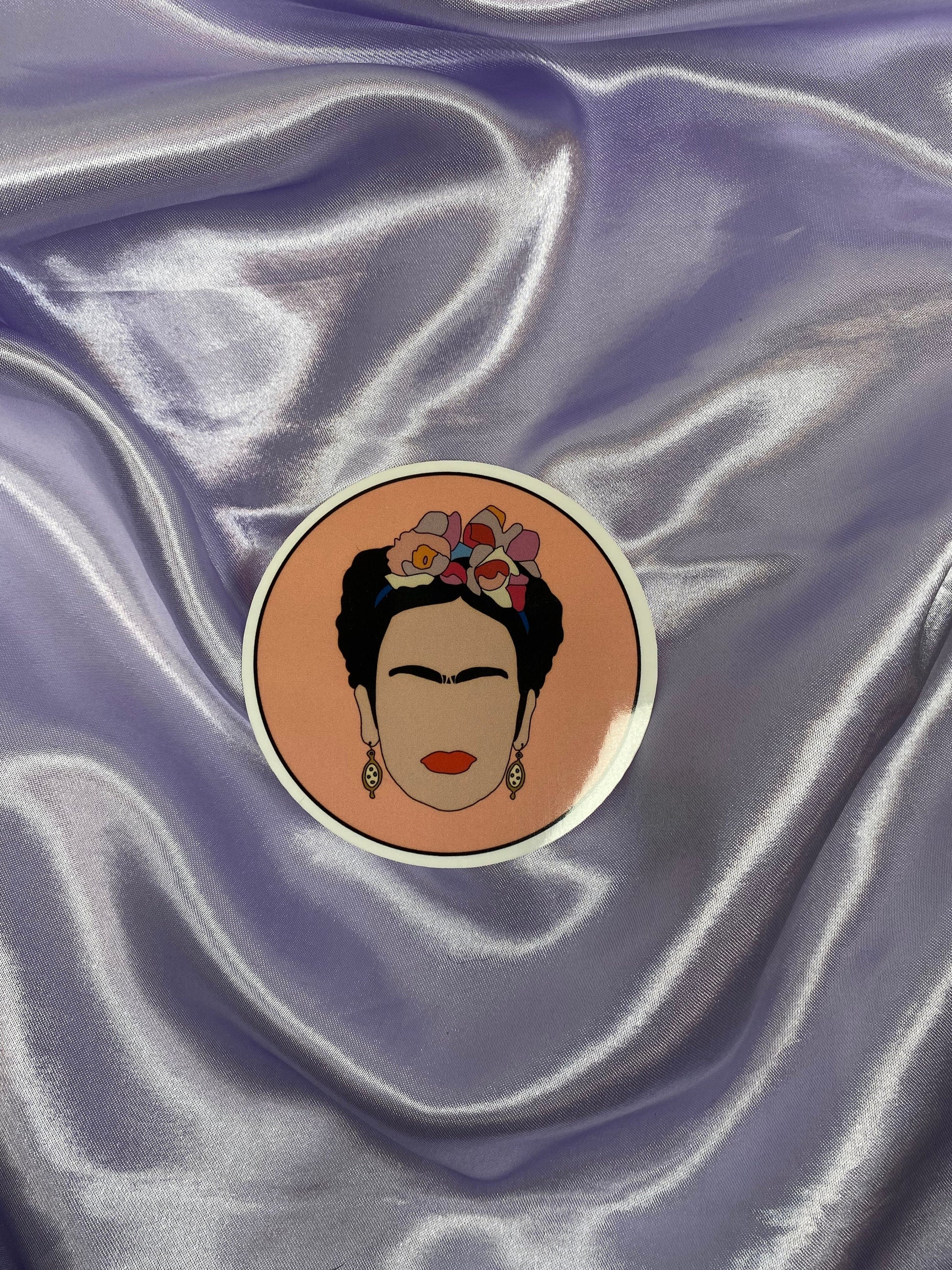 Circular sticker of a stylized portrait of Frida Kahlo on a textured purple fabric background