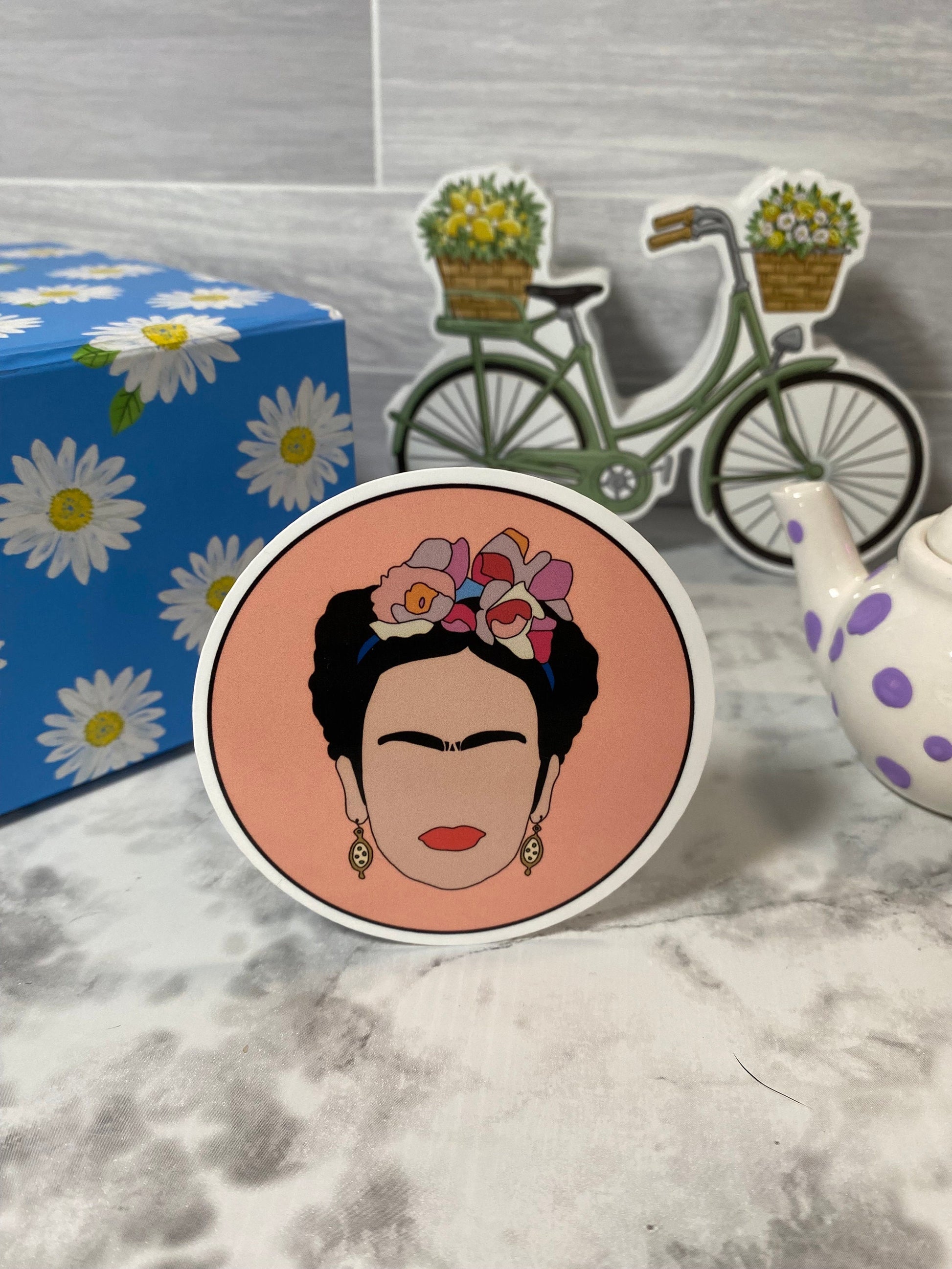 Circular sticker with a stylized portrait of Frida Kahlo with flowers in her hair, placed on a marble surface with a bicycle and box in the background.