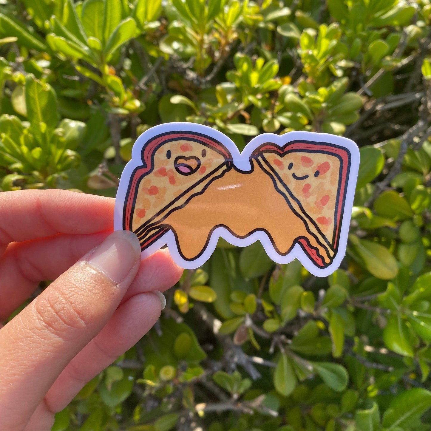 Hand holding a grilled cheese sticker with a smiley face against a green leafy background