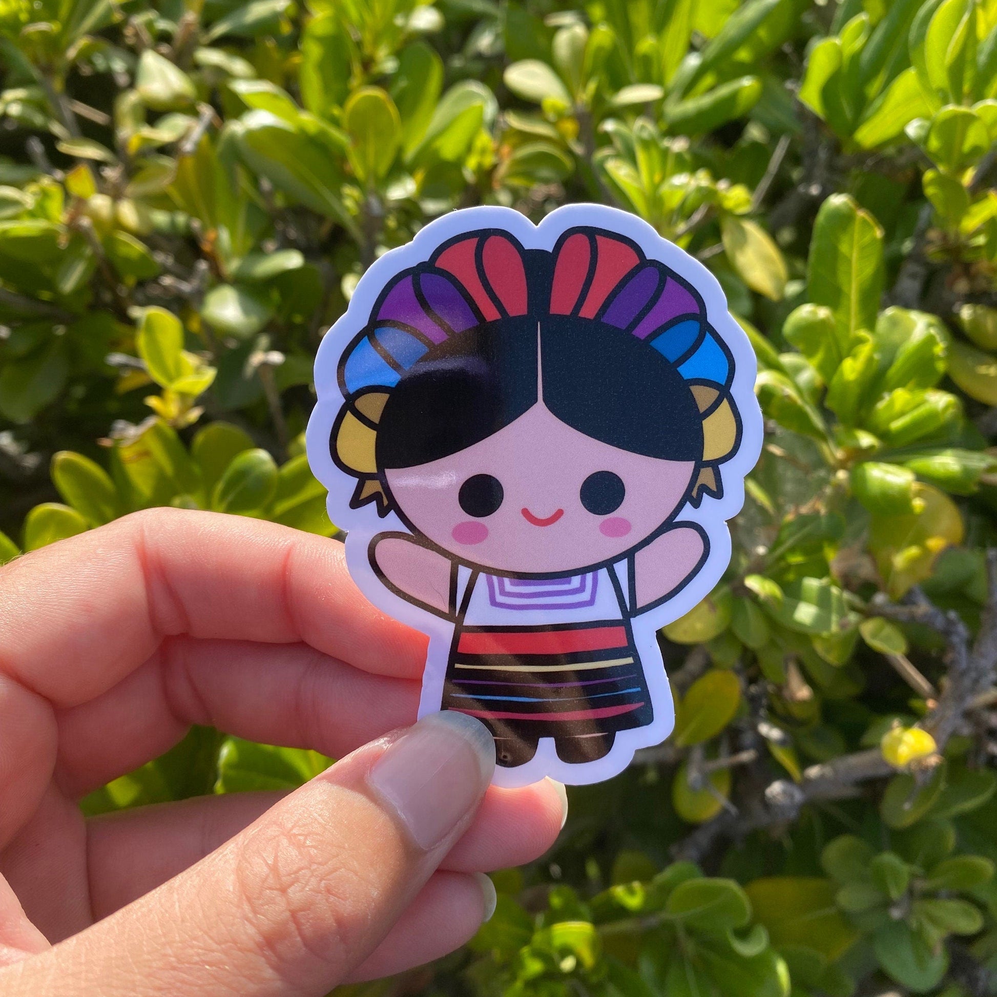 Hand holding a colorful sticker of a cartoon character with a rainbow headband against a green leafy background.