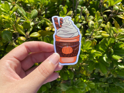 Hand holding a sticker of a cup with whipped cream against a green leafy background