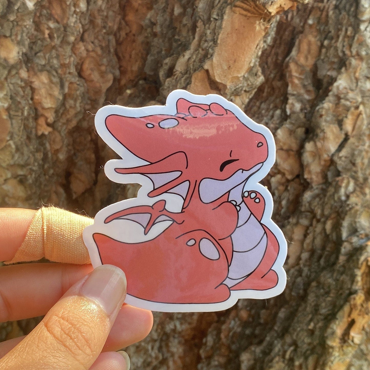 Hand holding a red dragon-shaped sticker against a tree bark background