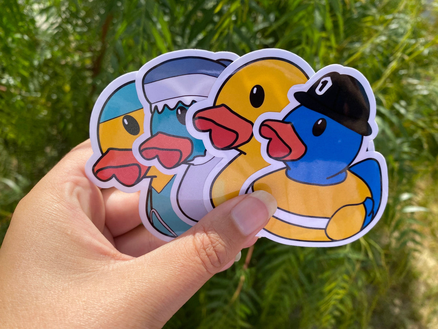 Four colorful duck-shaped stickers held by a hand with a green leafy background